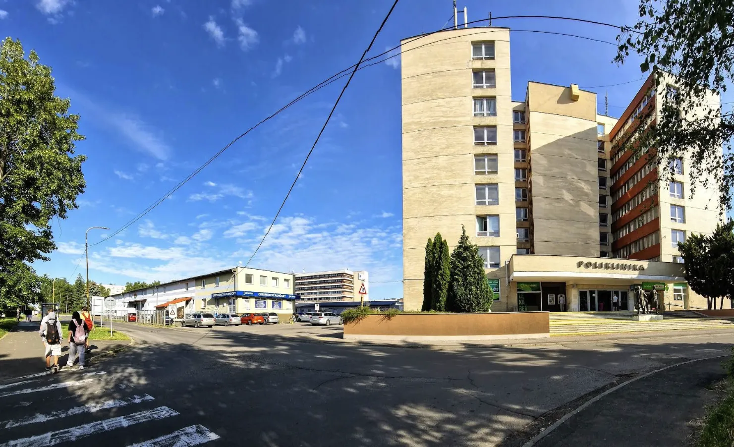 Budova s nápisom POLIKLINIKA, schody pri vchode a priľahlá ulica s prechodom pre chodcov v Humennom